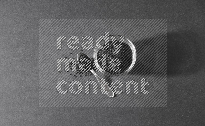 A glass bowl full of black seeds with a metal spoon full of the seeds on a black flooring