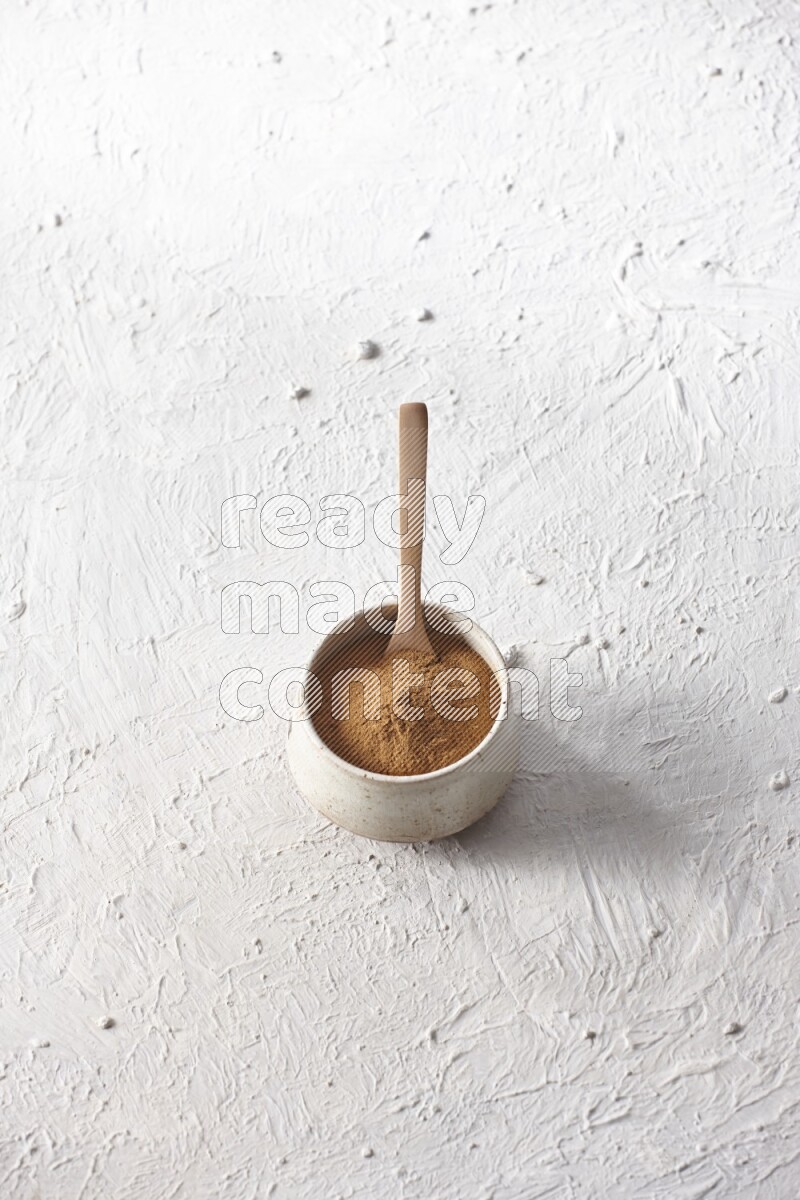Ceramic beige bowl full of cinnamon powder with a wooden spoon on a textured white background