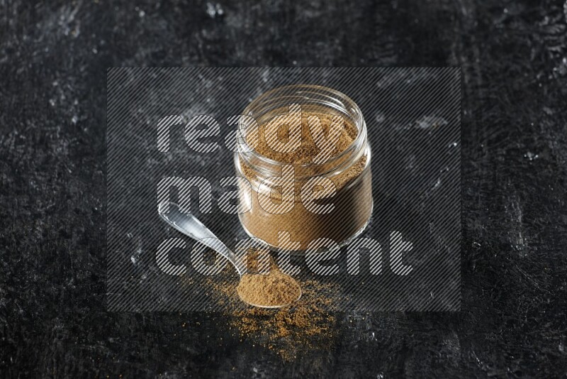 A glass jar and a metal spoon full of cumin powder on a textured black flooring