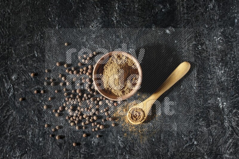 A wooden bowl and spoon full of allspice powder and whole balls spreaded on a black flooring