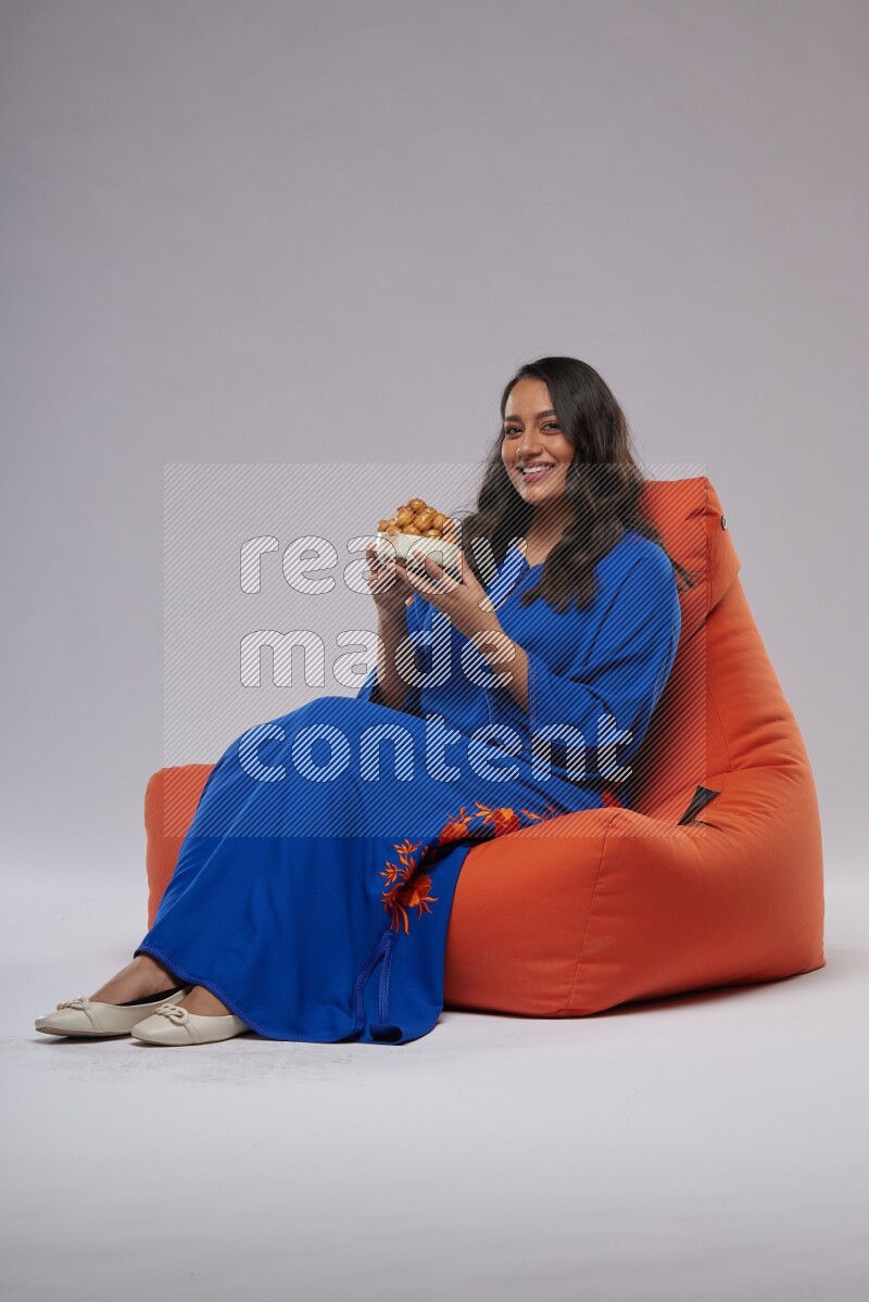 A Woman sitting on an orange beanbag wearing Jalabeya holding a plate of luqaimat