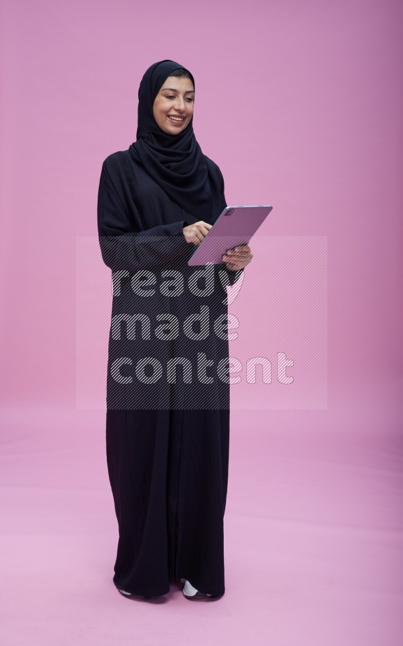 Saudi woman wearing Abaya standing working on tablet on pink background