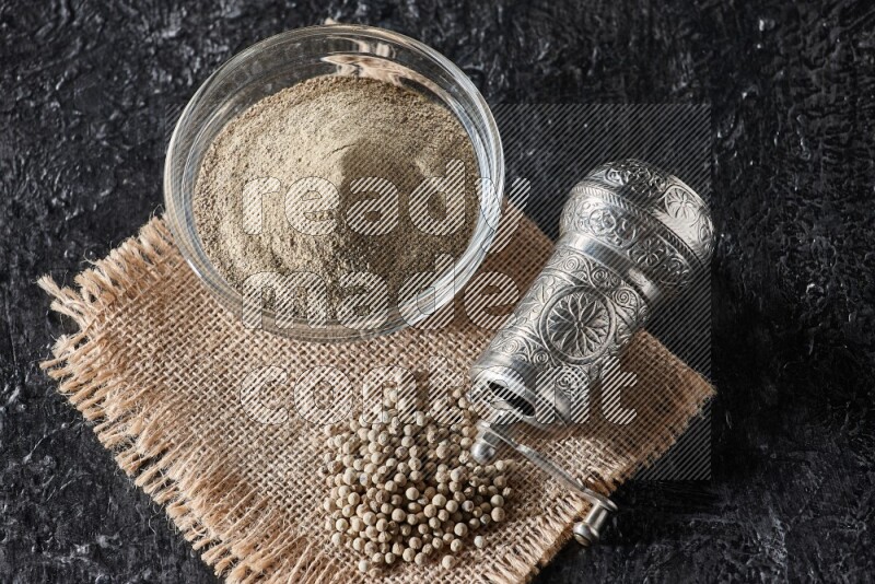 A glass bowl full of white pepper powder with white pepper beads on a burlap piece of fabric and a metal grinder on textured black flooring