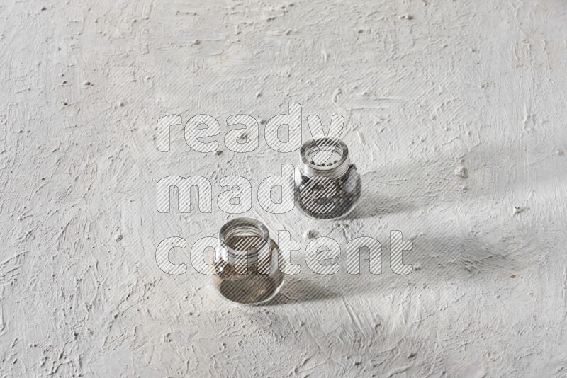 2 glass spice jars full of black pepper and black pepper powder on textured white flooring