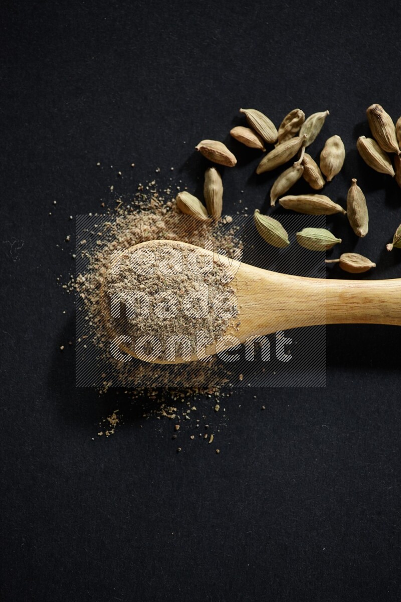 A wooden spoon full of cardamom powder and cardamom seeds beside it on black flooring
