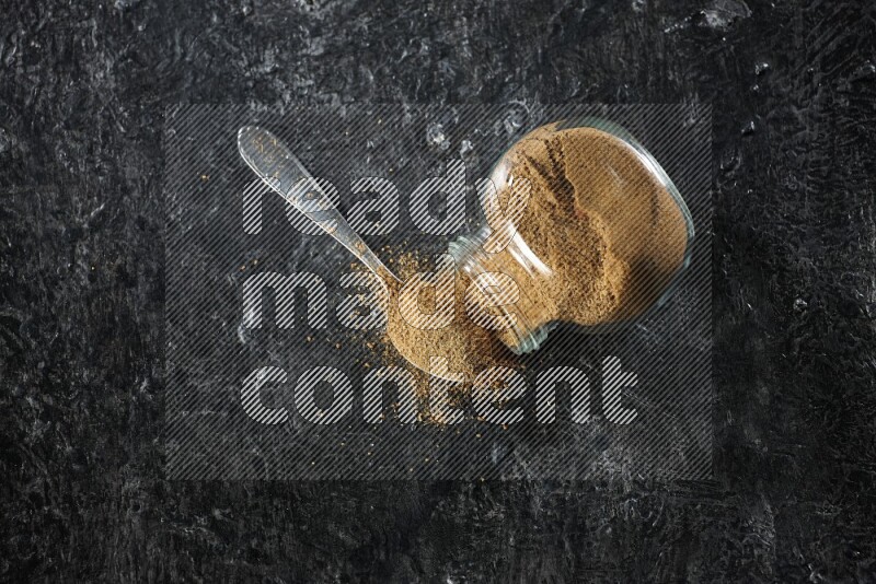 وعاء زجاجي للتوابل مقلوب وملعقة معدنية ممتلئة ببودرة الكمون والبودرة تناثرت منه على خلفية سوداء