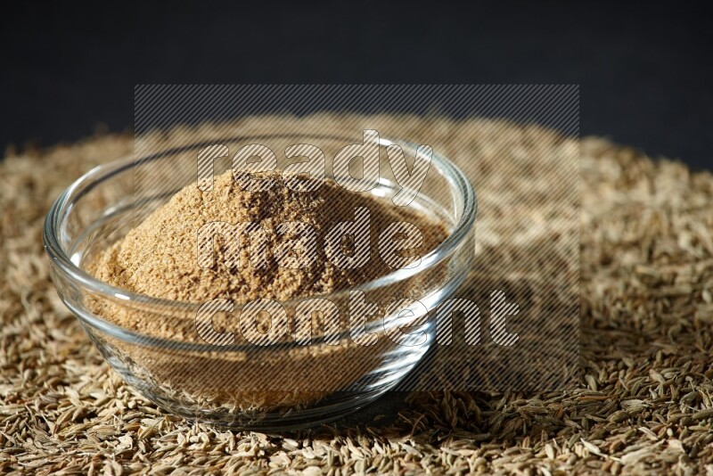 A glass bowl full of cumin powder surrounded by cumin seeds on black flooring