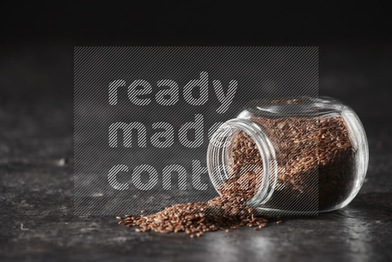 A glass spice jar full of flaxseeds flipped and seeds spread out on a textured black flooring