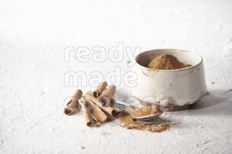 Ceramic beige bowl full of cinnamon powder and a metal spoon with cinnamon sticks next of it on a textured white background