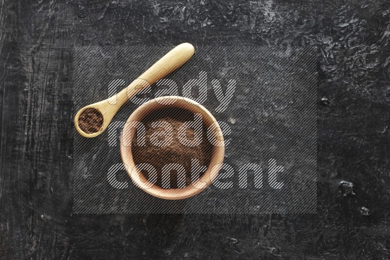 A wooden bowl and wooden spoon full of cloves powder on textured black flooring