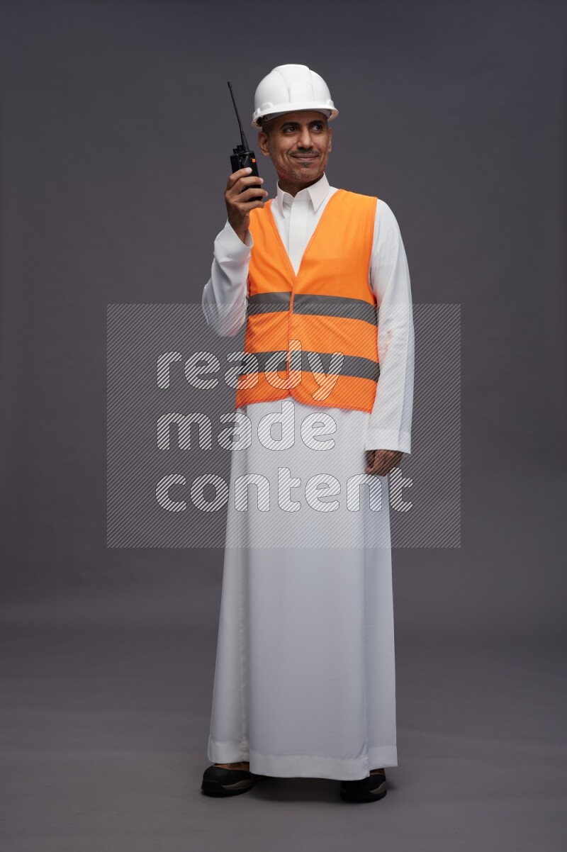 Saudi man wearing thob with engineer vest standing holding walkie-talkie on gray background