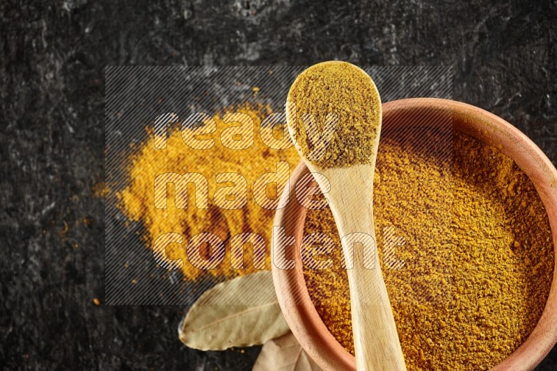 A wooden bowl and a wooden spoon full of turmeric powder on textured black flooring