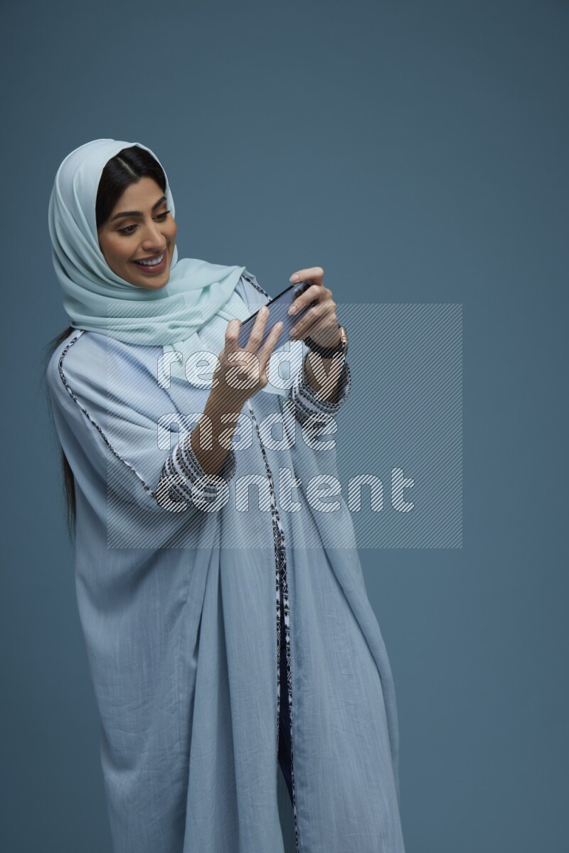 A Saudi woman Playing a Game on her phone on a blue background wearing a blue Abaya with hijab
