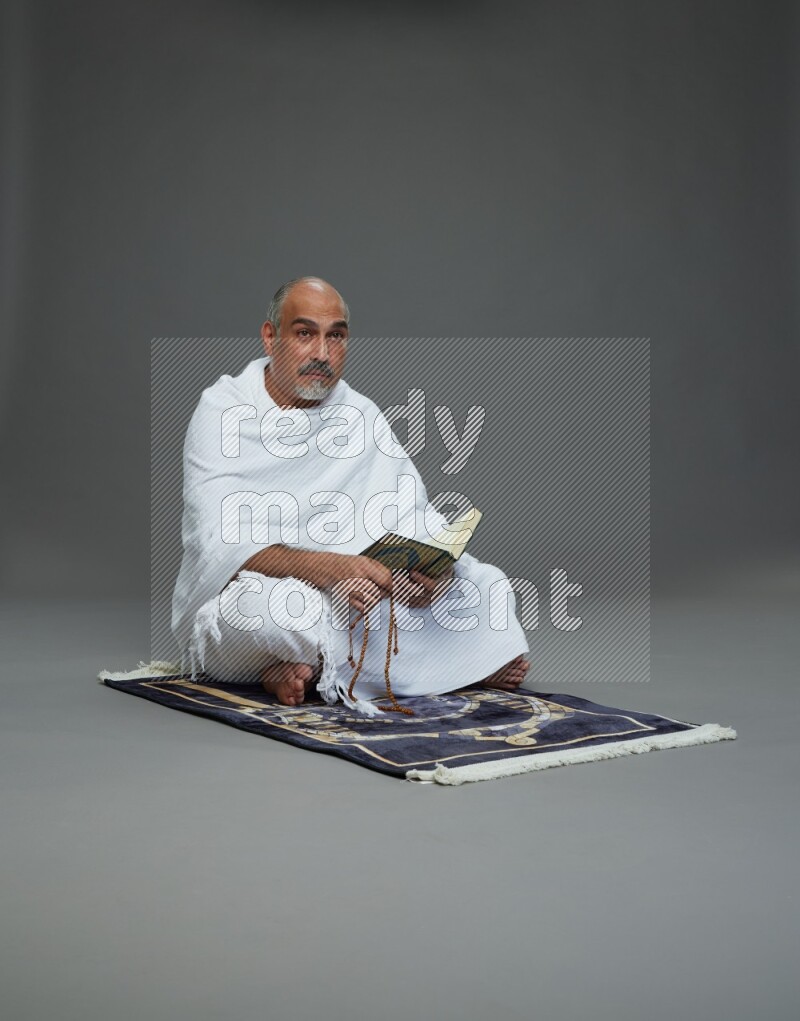 A man wearing Ehram sitting on mate prayer reading quran on gray background