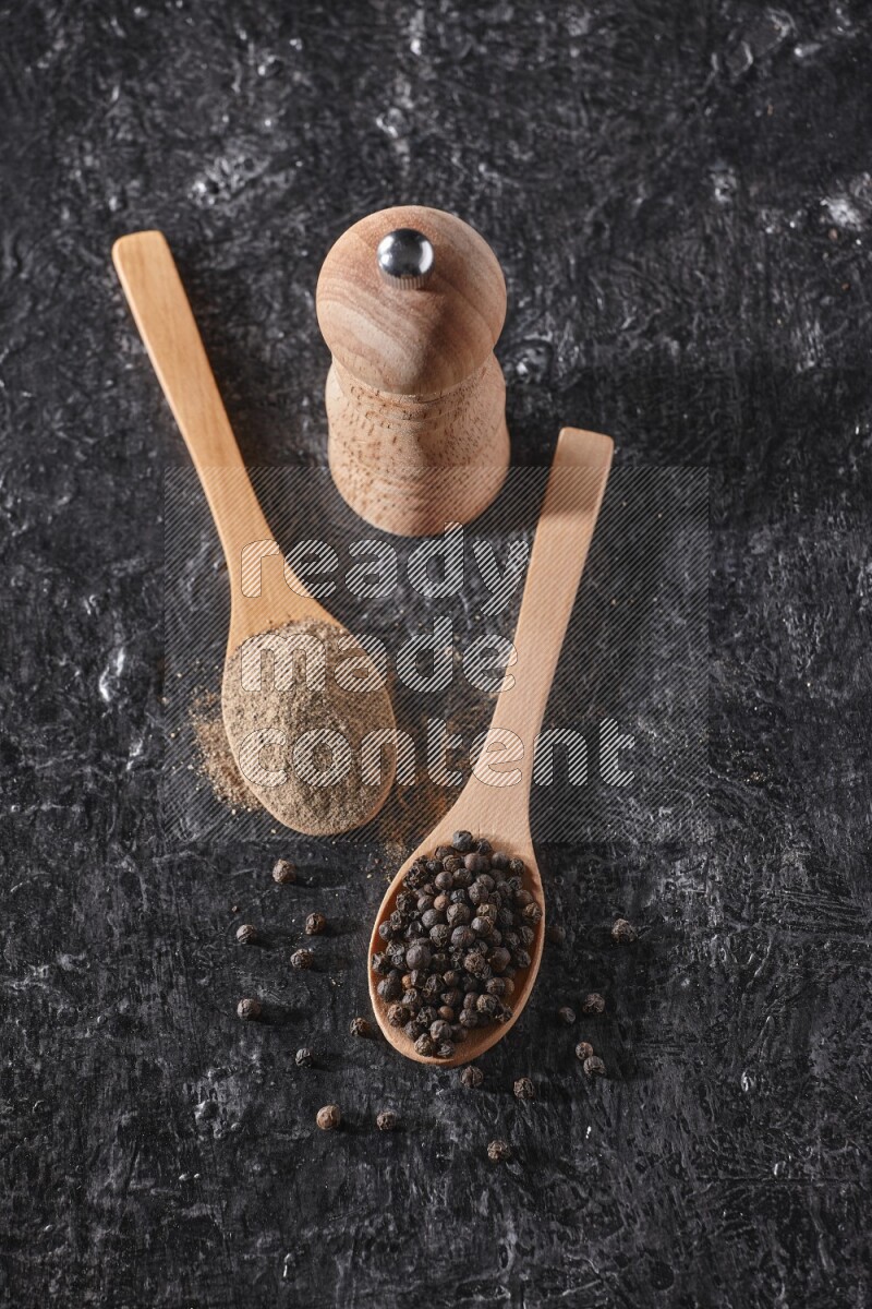 Black pepper powder and black pepper beads in wooden spoons and wooden grinder on a textured black flooring