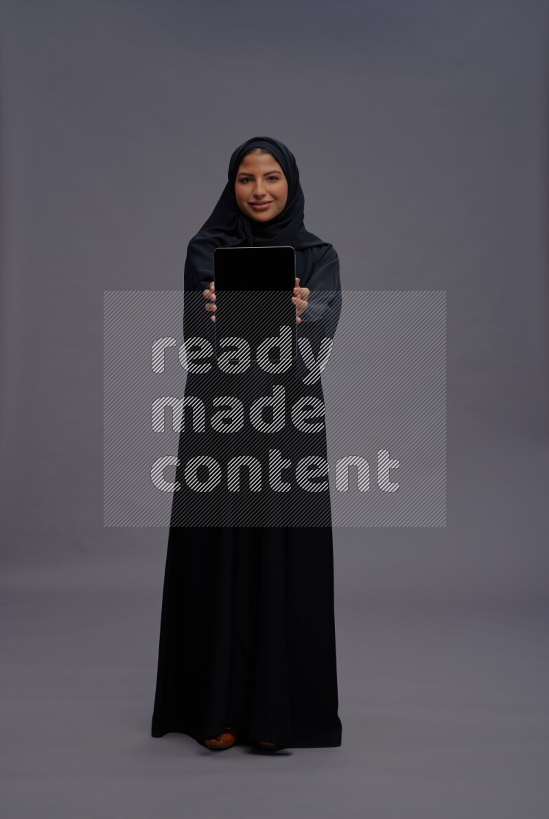 Saudi woman wearing Abaya standing showing tablet to camera on gray background