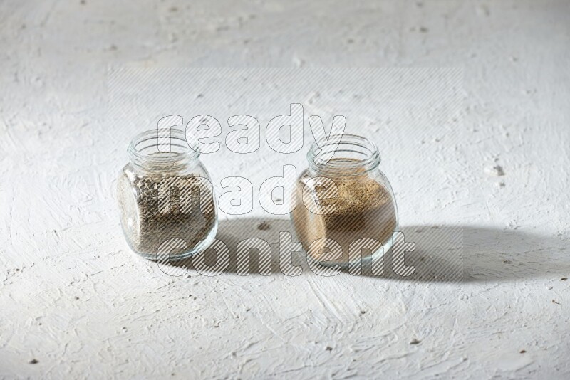 2 glass spice jars full of cumin powder and cumin seeds on textured white flooring