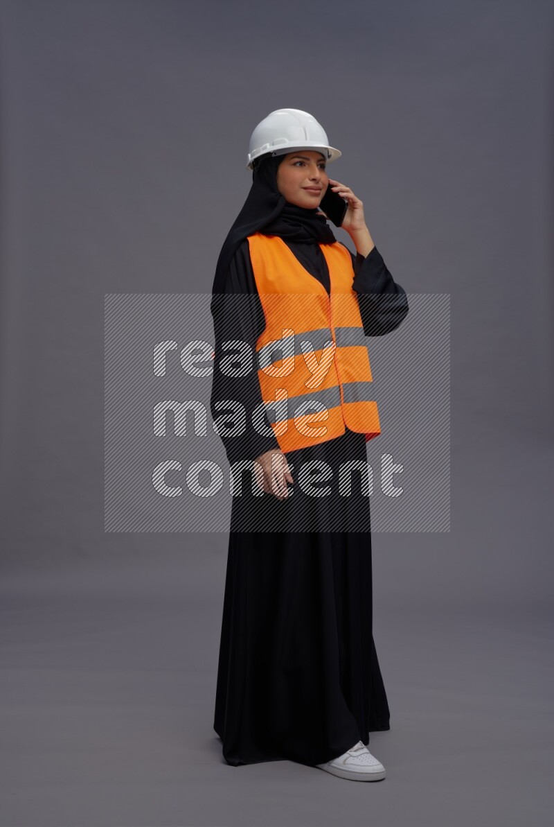 Saudi woman wearing Abaya with engineer vest standing talking on phone on gray background