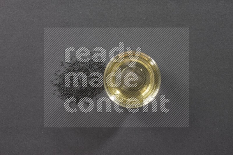 A glass bowl full of black seeds oil and black seeds beside it on a black flooring