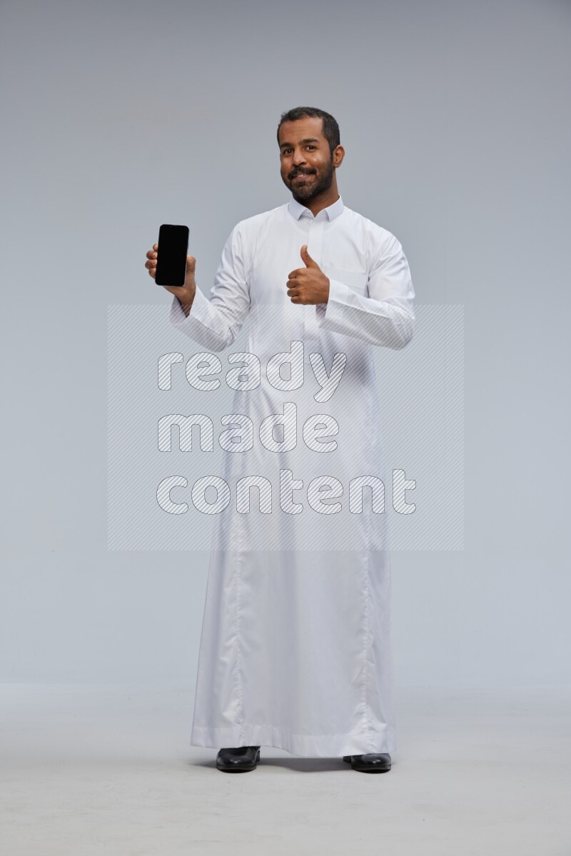 Saudi man wearing Thob standing showing phone to camera on Gray background