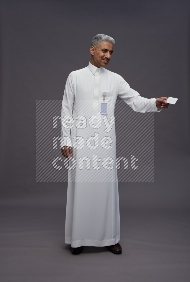 Saudi man wearing thob with pocket employee badge standing holding ATM card on gray background