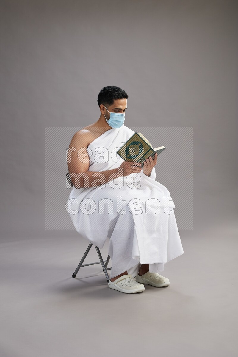 A man wearing Ehram with face mask sitting on chair reading quran on gray background
