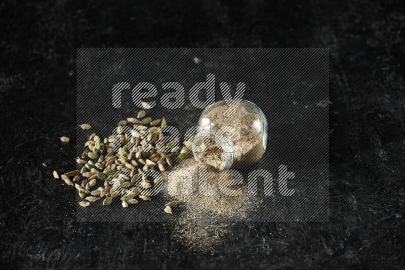 A flipped glass spice jar full of cardamom powder and powder spilled out of it with cardamom seeds spreaded on textured black flooring