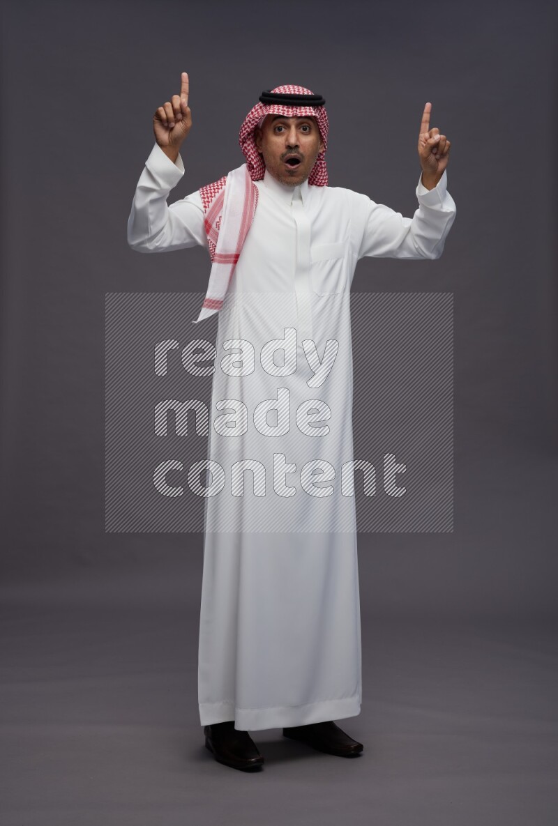 Saudi man wearing thob and shomag standing interacting with the camera on gray background