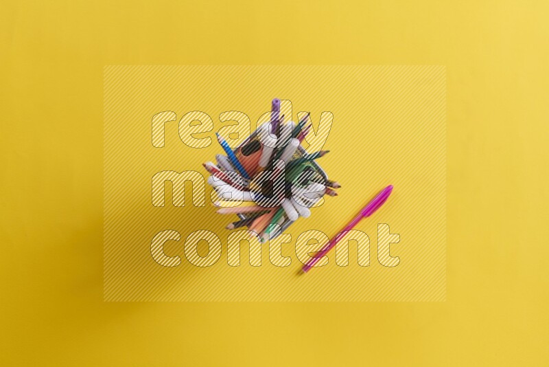 Pens, pencils and school supplies in a metal pen holder on yellow background