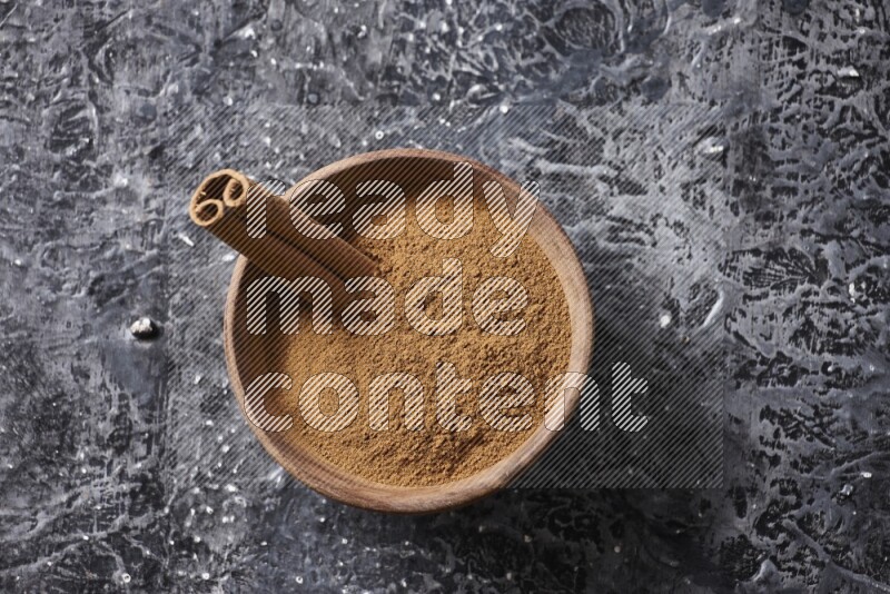 Wooden bowl full of cinnamon powder and a cinnamon stick on a textured black background