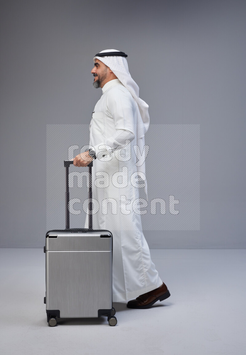 Saudi man wearing Thob and white Shomag standing holding Travel bag on Gray background