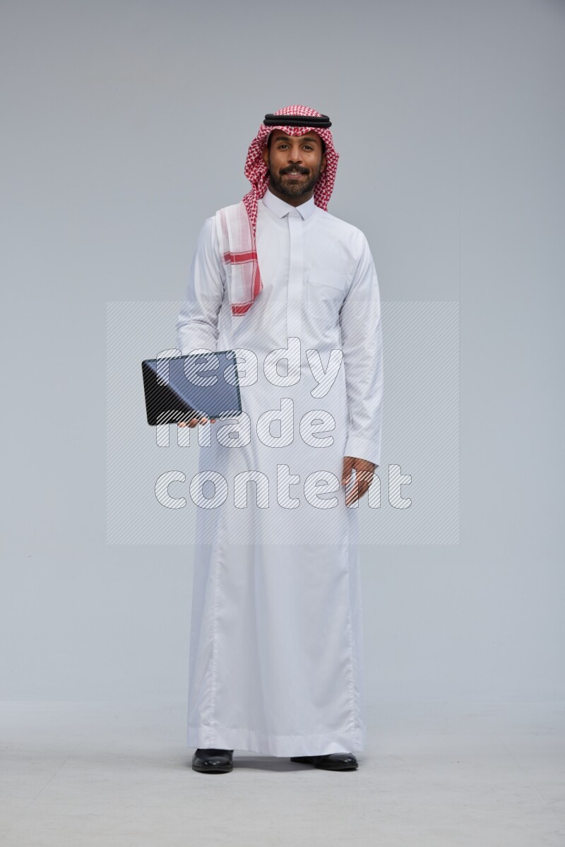 Saudi man Wearing Thob and shomag standing showing tablet to camera on Gray background