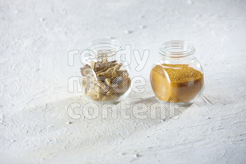 2 glass spice jars full of turmeric powder and dried whole fingers on textured white flooring