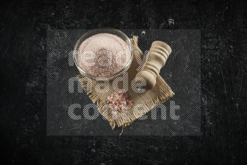 A glass bowl full of pink himalayan salt with a wooden grinder on a burlap fabric all on black background