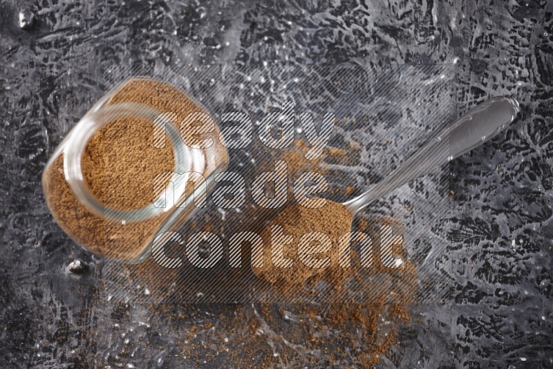 Herbal glass jar full of cinnamon powder and a metal spoon full of powder on textured black background