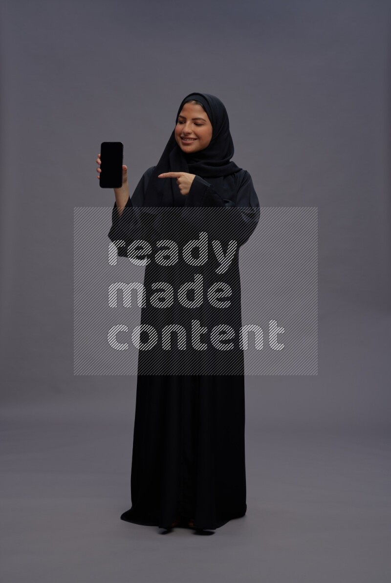 Saudi woman wearing Abaya standing showing phone to camera on gray background