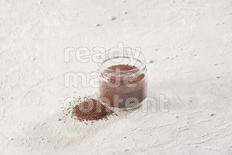 A glass jar full of garden cress seeds with more seeds spread on a textured white flooring