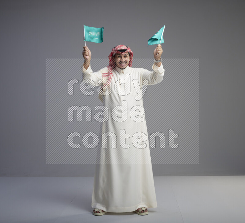 A Saudi man standing wearing thob and red shomag raising small Saudi flag on gray background
