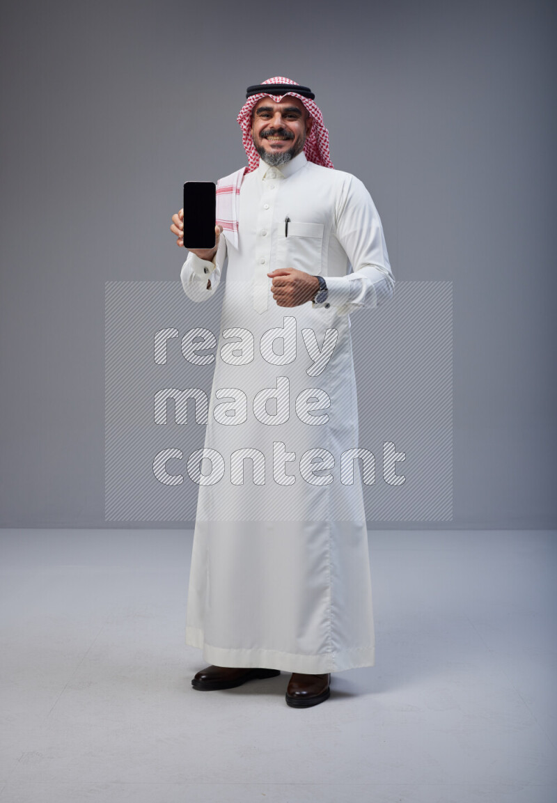 Saudi man Wearing Thob and red Shomag standing showing phone to camera on Gray background