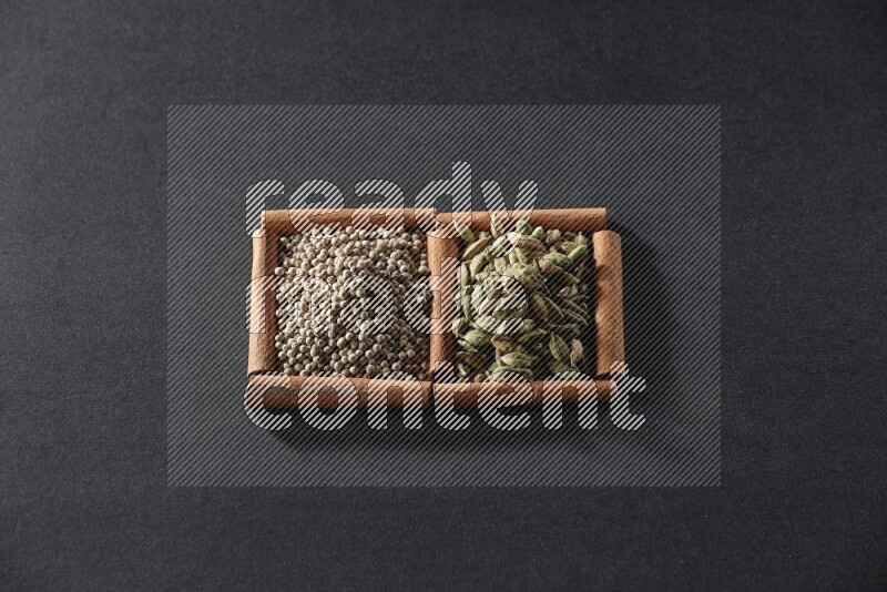 مربعان من أعواد القرفة ممتلئان بحبوب الفلفل الأبيض وحبوب الهيل علي خلفية سوداء