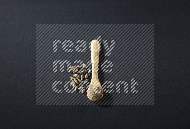 A wooden spoon full of cardamom powder and cardamom seeds beside it on black flooring