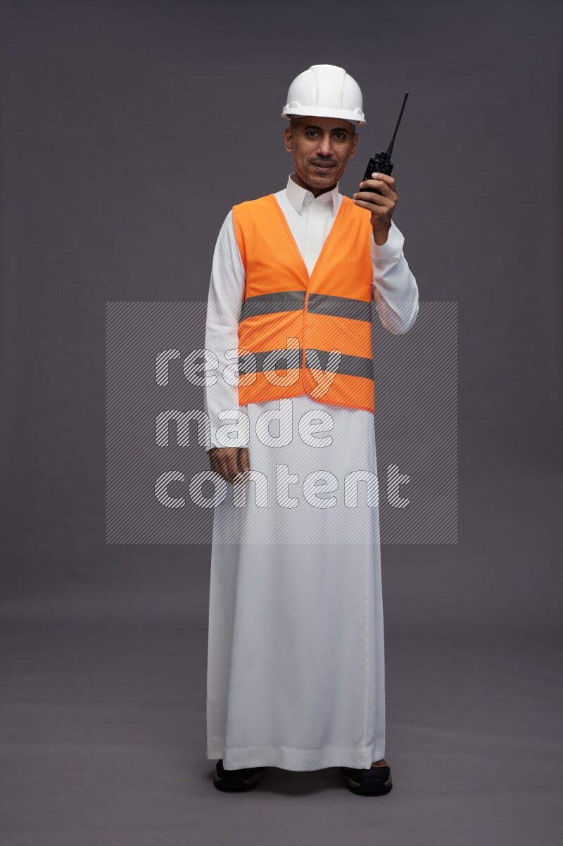 Saudi man wearing thob with engineer vest standing holding walkie-talkie on gray background