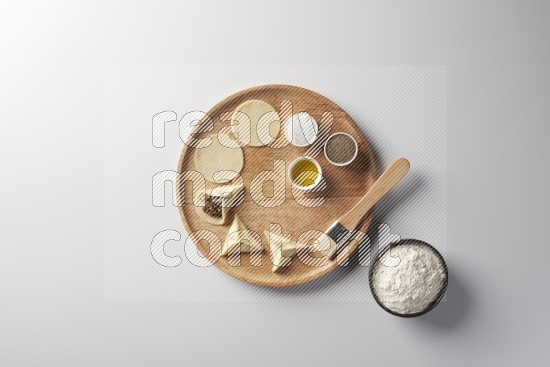 two closed sambosas and one open sambosa filled with meat while flour, salt, black pepper and oil with oil brush aside in a wooden dish on a white background