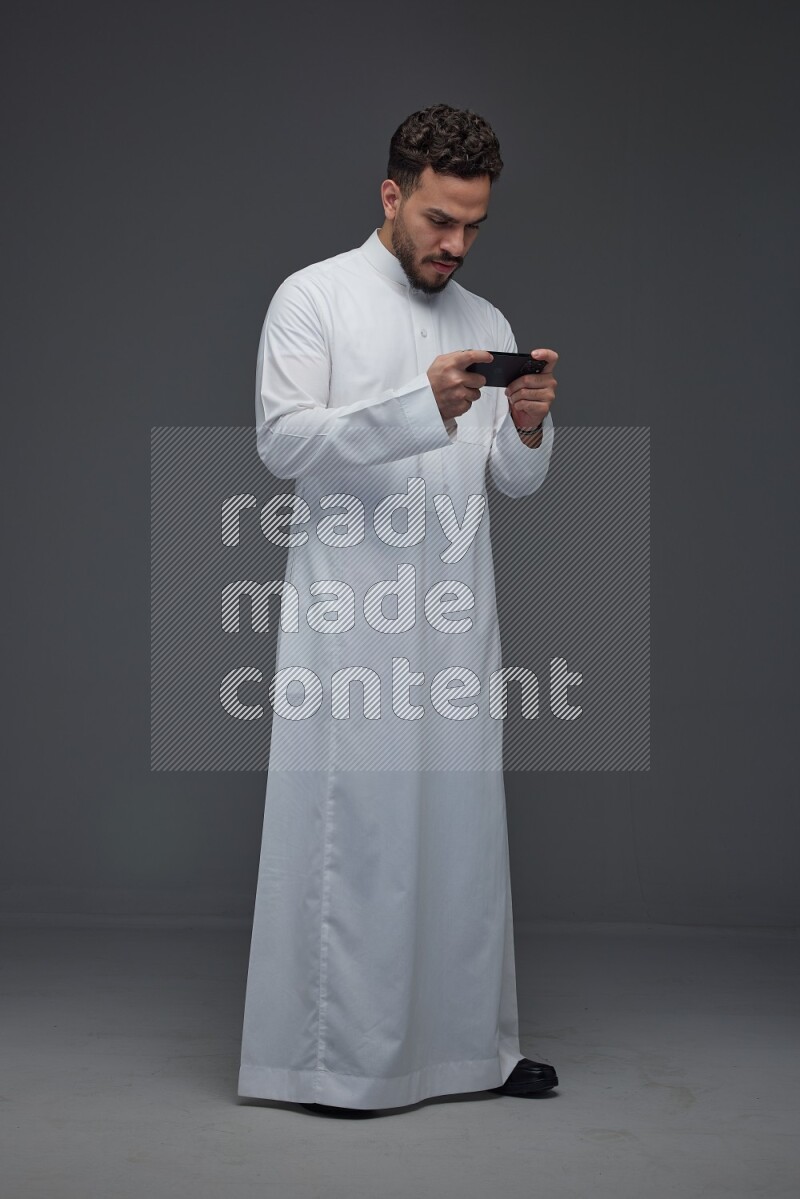 A Saudi man wearing Thobe and playing using his phone horizontally playing video games while standing and making different poses eye level on a gray background