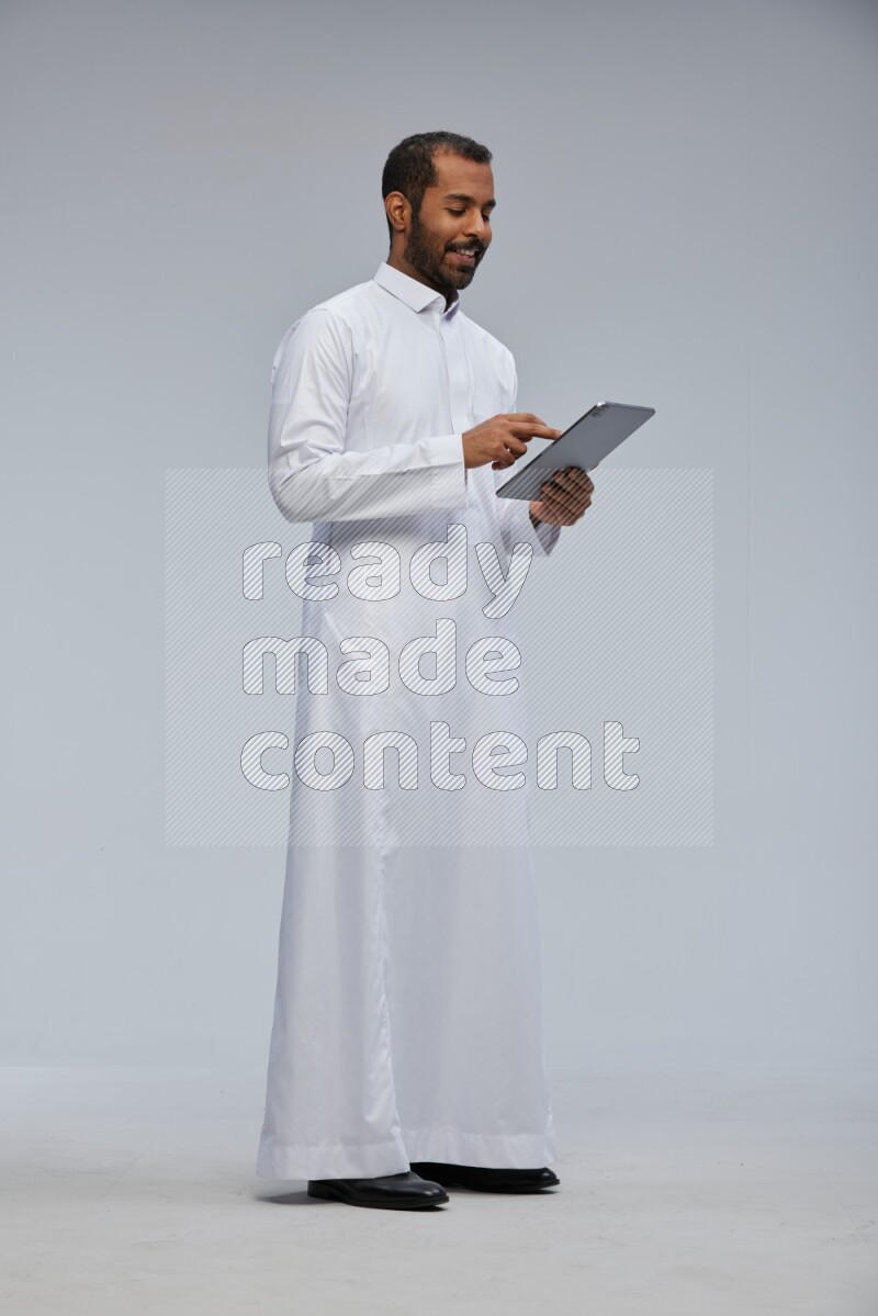 Saudi man wearing Thob standing working on tablet on Gray background