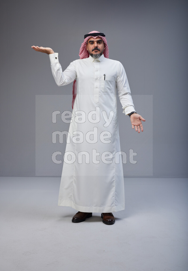 Saudi man Wearing Thob and red white Shomag standing interacting with the camera on Gray background