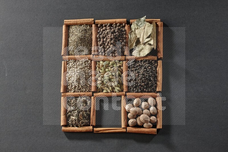 9 squares of cinnamon sticks full of cardamom in the middle surrounded by nutmeg, black pepper, bay laurel leaves, allspice, cumin, white pepper, dried basil and salt on black flooring
