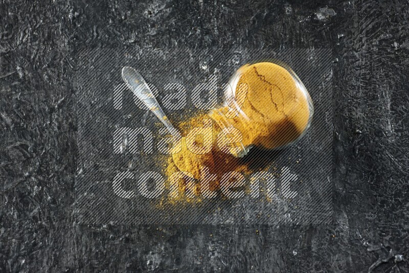 A flipped glass spice jar and a metal spoon full of turmeric powder and powder spilled out of it on textured black flooring