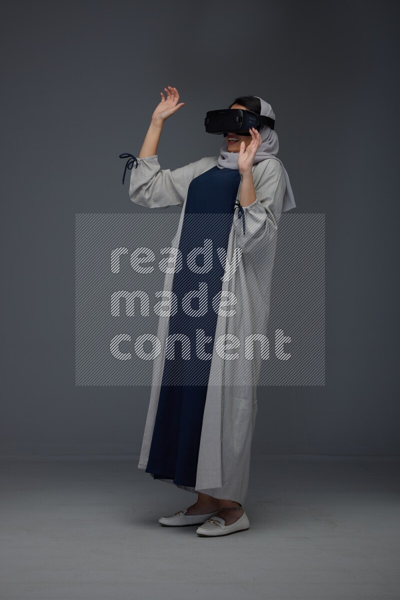 A Saudi woman wearing a light gray Abaya and head scarf standing and wearing VR eye level on a grey background