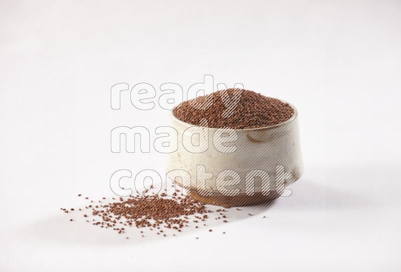 A beige pottery bowl full of garden cress seeds with more seeds spread on a white flooring